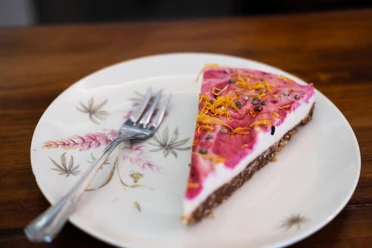 Vevi Ein Stück Kuchen mit pinker Glasur, bestreut mit Blüten, liegt auf einem floral verzierten Teller.