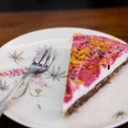 Vevi Ein Stück Kuchen mit pinker Glasur, bestreut mit Blüten, liegt auf einem floral verzierten Teller.A piece of cake with pink icing, sprinkled with flowers, lies on a plate decorated with floral motifs.