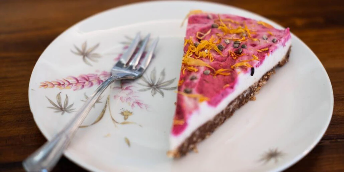 Vevi Ein Stück Kuchen mit pinker Glasur, bestreut mit Blüten, liegt auf einem floral verzierten Teller.A piece of cake with pink icing, sprinkled with flowers, lies on a plate decorated with floral motifs.
