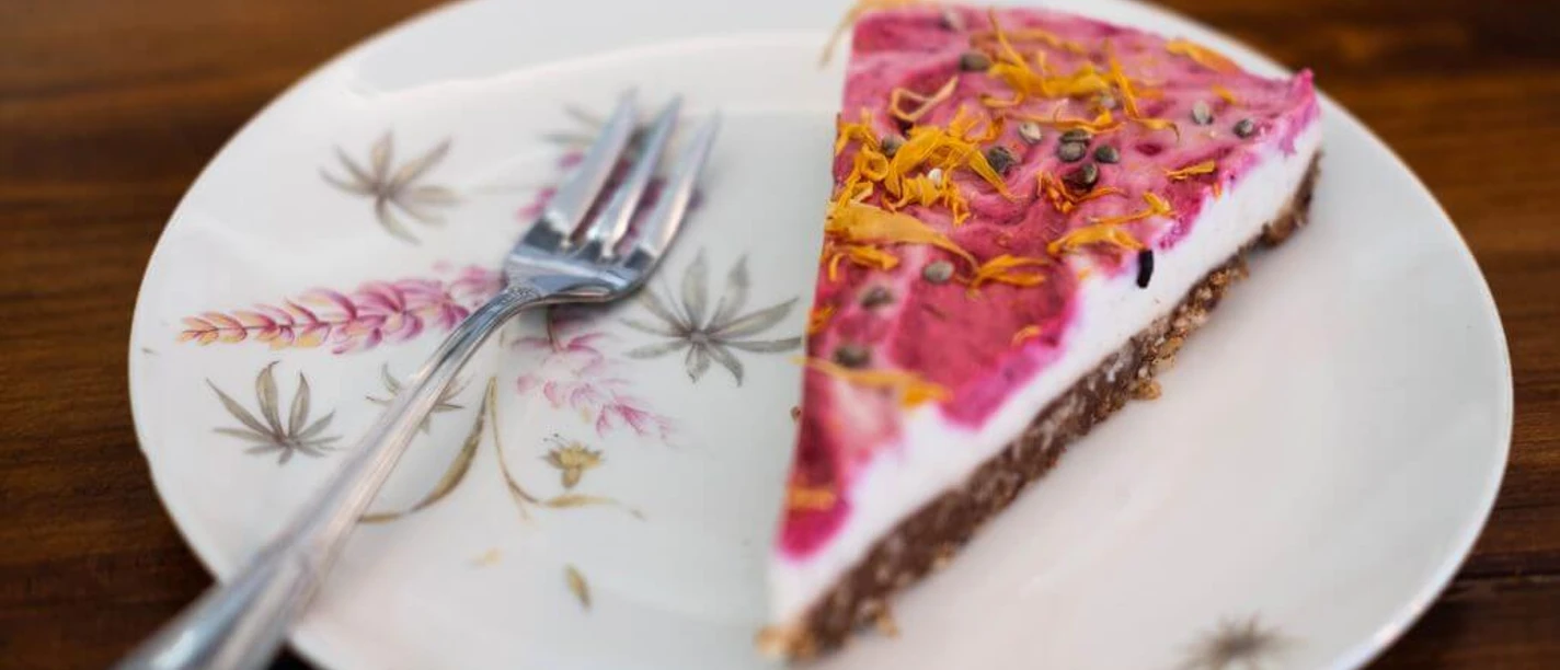 Vevi Ein Stück Kuchen mit pinker Glasur, bestreut mit Blüten, liegt auf einem floral verzierten Teller.