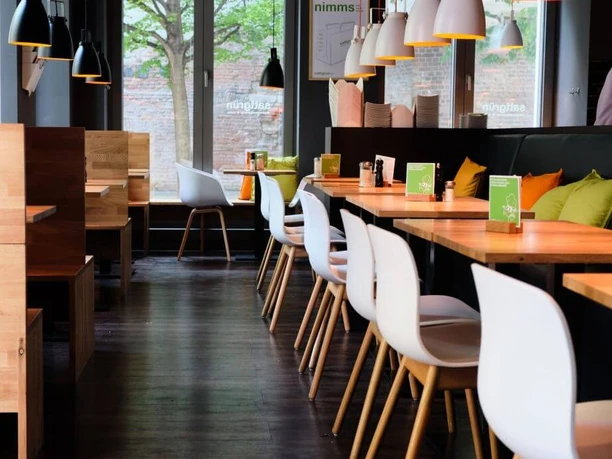 Sattgrün In a stylishly decorated restaurant with contemporary lighting, wooden tables are arranged in rows, complemented by white designer chairs. The windows offer green views, while small cards displaying menu offerings are placed on the tables.