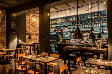 Phaedra A stylishly furnished restaurant with a cosy atmosphere, exposed brick walls, and thoughtfully arranged wines displayed on shelves. Bright lighting accentuates the bar area, while wooden tables and chairs invite guests to linger.