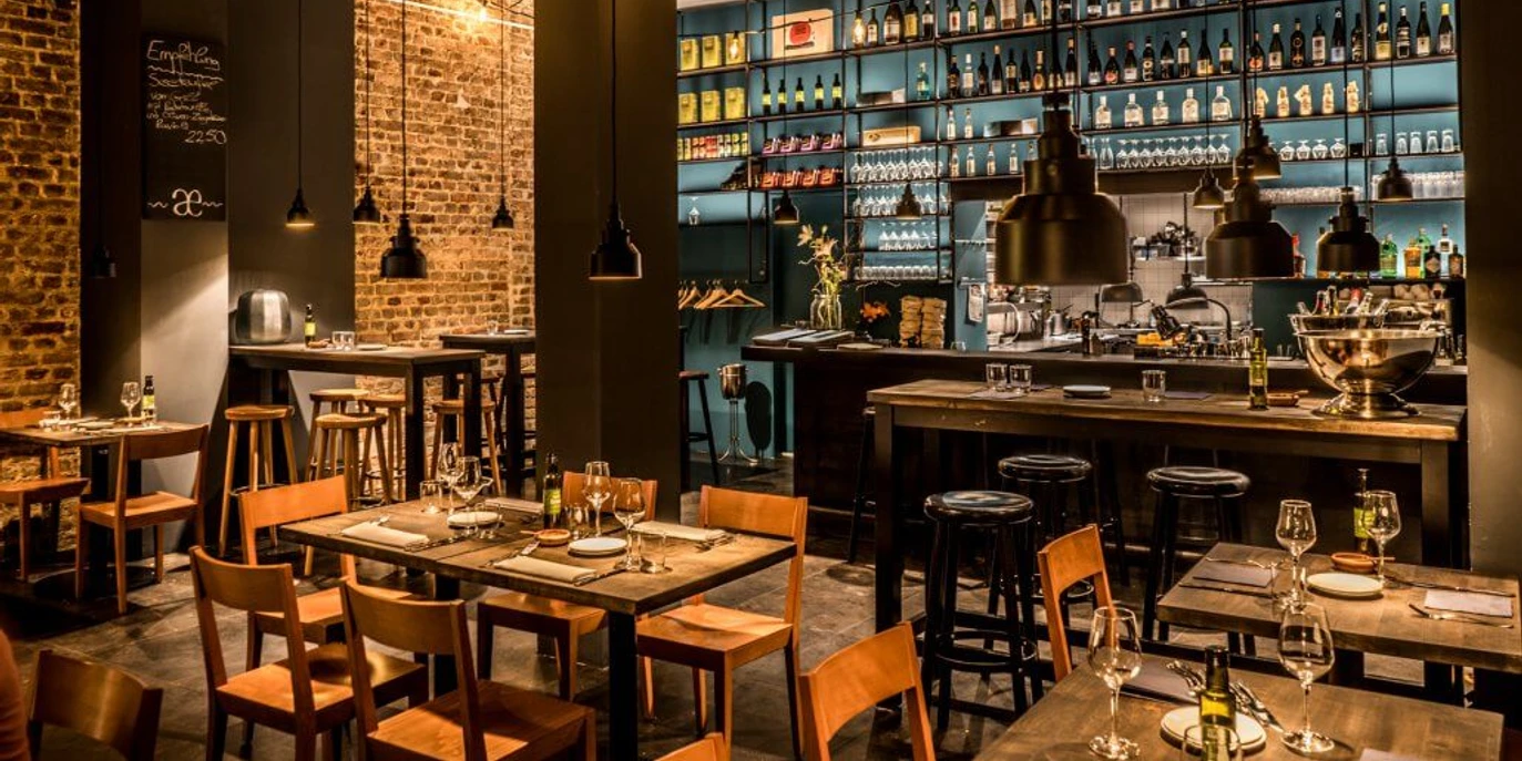 Phaedra Ein stilvoll eingerichtetes Restaurant mit gemütlicher Atmosphäre, freiliegenden Backsteinwänden und sorgfältig arrangierten Weinen in Regalen. Helle Beleuchtung akzentuiert den Barbereich, während Holztische und Stühle zum Verweilen einladen.A stylishly furnished restaurant with a cosy atmosphere, exposed brick walls, and thoughtfully arranged wines displayed on shelves. Bright lighting accentuates the bar area, while wooden tables and chairs invite guests to linger.