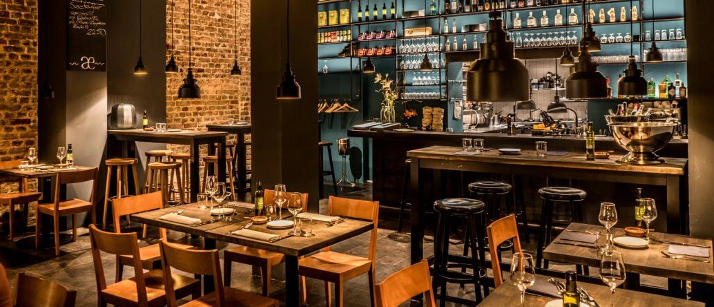 Phaedra A stylishly furnished restaurant with a cosy atmosphere, exposed brick walls, and thoughtfully arranged wines displayed on shelves. Bright lighting accentuates the bar area, while wooden tables and chairs invite guests to linger.