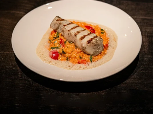 Phaedra Ein perfekt gegrilltes Fischfilet auf einem Bett aus cremigem Risotto, dekoriert mit frischen Kirschtomaten und Duft von frischem Basilikum.A perfectly grilled fish fillet served on a bed of creamy risotto, garnished with fresh cherry tomatoes and scented with aromatic basil.