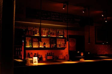 Pegel Auf dem Bild zeigt sich eine stimmungsvoll beleuchtete Bar mit einer Reihe von Flaschen und Gläsern hinter dem Tresen. Warme Lichter schaffen eine einladende Atmosphäre.