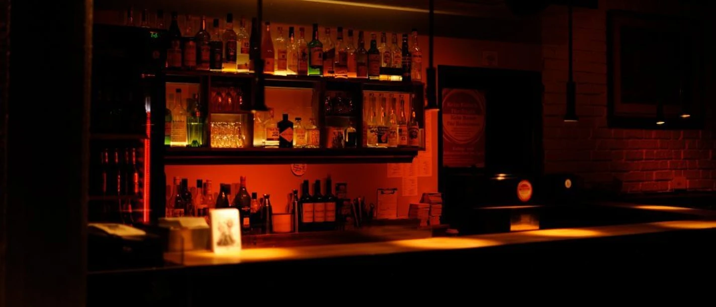 Pegel The picture shows an atmospherically lit bar with a row of bottles and glasses behind the counter. Warm lights create an inviting atmosphere.