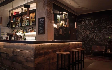Bar em Schnörres Holzverkleidete Bartheke mit Hockern in schummrigem Licht. Eine Kreidetafel ziert die komplette hintere Wand mit Kritzeleien drauf.