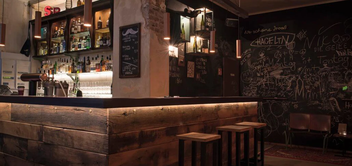 Bar em Schnörres Holzverkleidete Bartheke mit Hockern in schummrigem Licht. Eine Kreidetafel ziert die komplette hintere Wand mit Kritzeleien drauf.Wood-paneled bar with stools in dim light. A chalkboard adorns the entire back wall with scribbles on it.