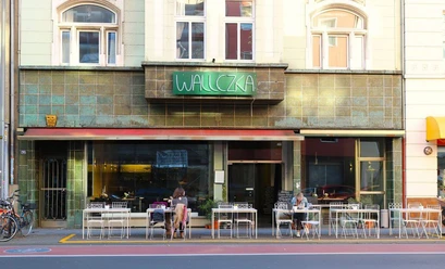 Wallczka The street scene depicts the restaurant Wallczka with large windows, two guests, and a green neon sign.
