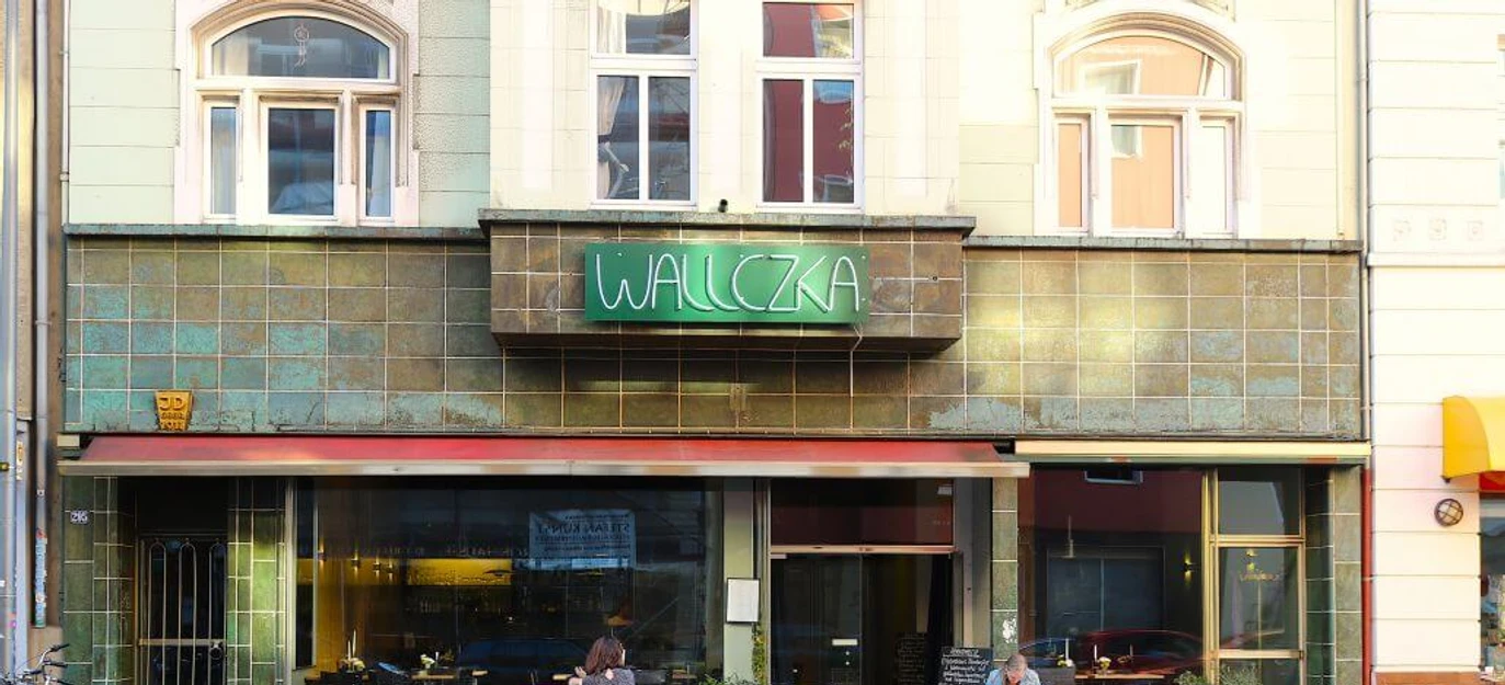 Wallczka Die Straßenszene zeigt das Restaurant Wallczka mit großen Fenstern, zwei Gästen und einem grünen Neonschild.The street scene depicts the restaurant Wallczka with large windows, two guests, and a green neon sign.
