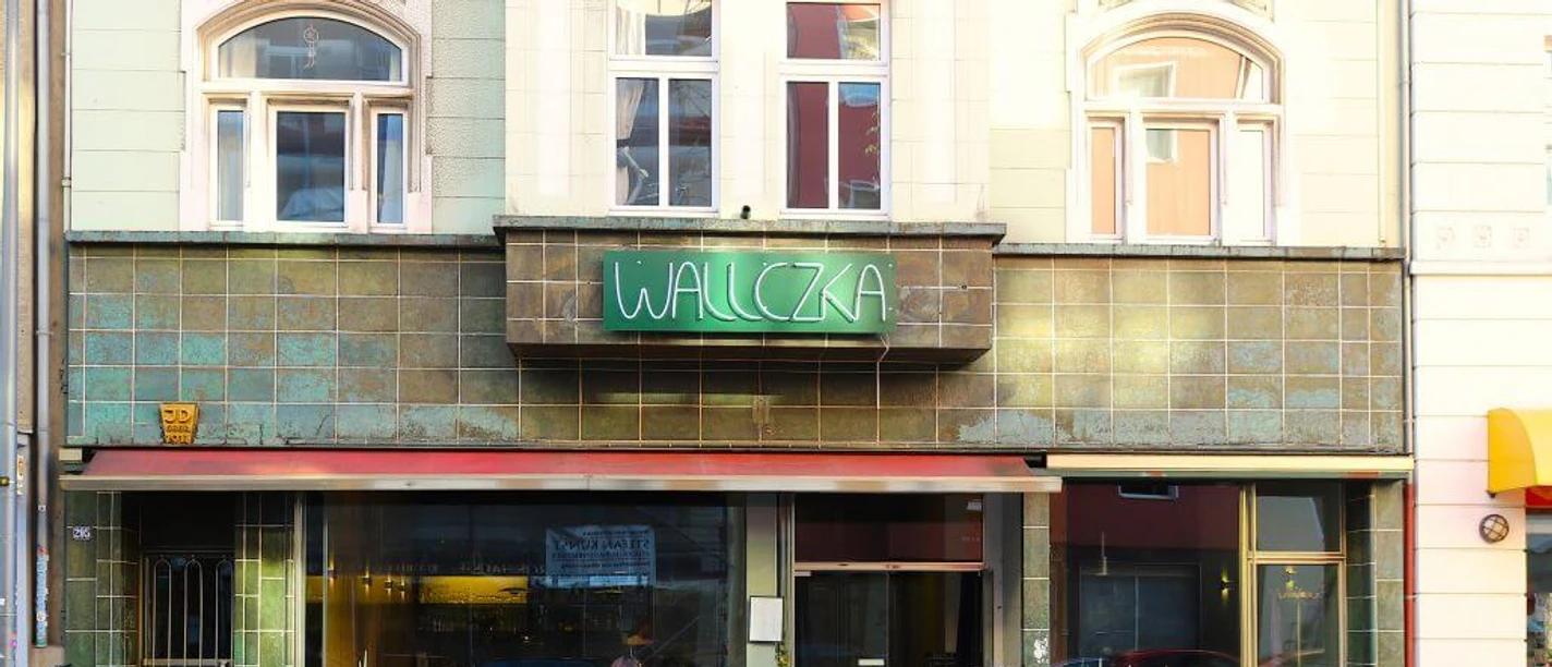 Wallczka Die Straßenszene zeigt das Restaurant Wallczka mit großen Fenstern, zwei Gästen und einem grünen Neonschild.