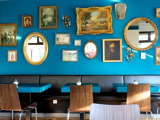 Cafe Jealys Innenansicht im Restaurant, Gemälde in goldenen Rahmen hängen an einer blauen Wand. Davor stehen Tische mit Stühlen.Interior view of a restaurant, paintings in golden frames hanging on a blue wall. In front of it are tables with chairs.
