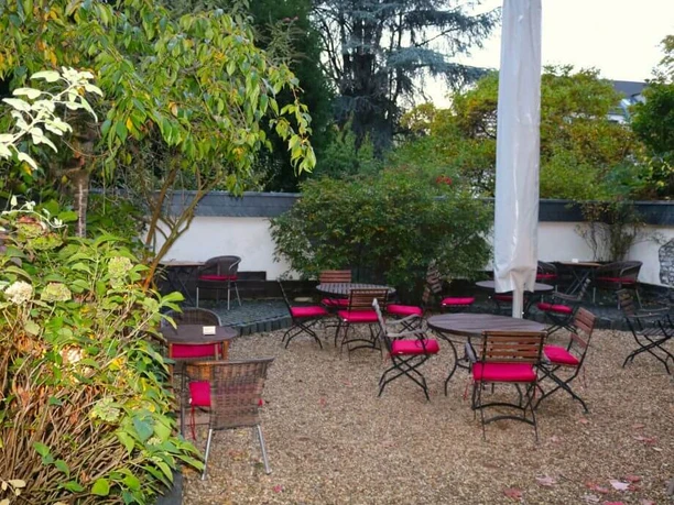 Traubenzeit A charming summer garden featuring red chairs, round tables, and bordered by sun umbrellas, all placed on a gravel surface.