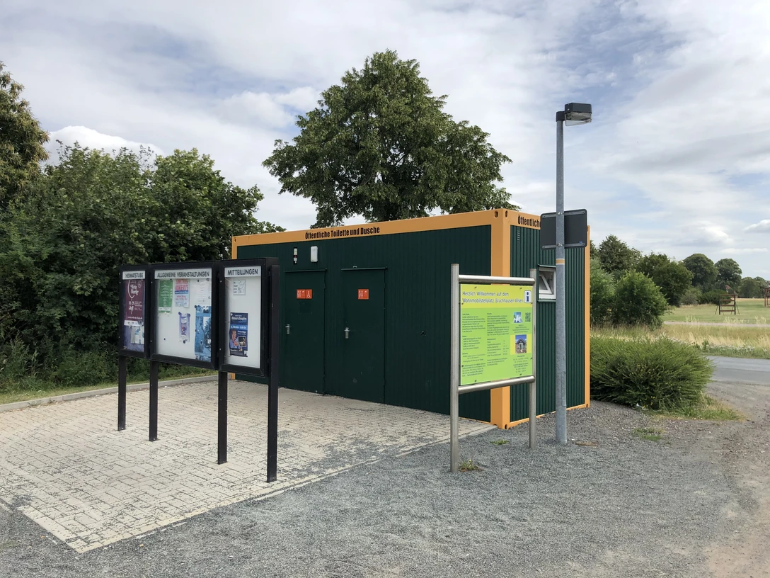 Grüne Sanitäranlage an einem Wohnmobilstellplatz mit Informationsschild und gepflastertem Vorplatz.