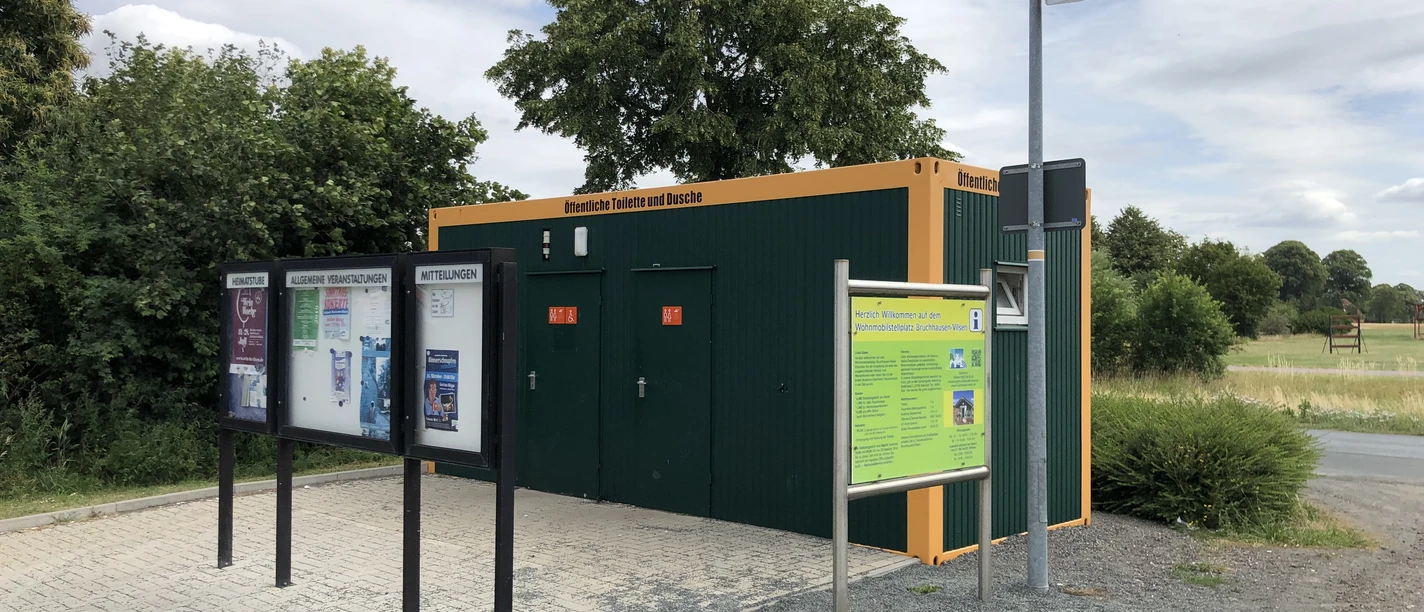 Toilette am Wohnmobilstellplatz Bruchhausen-Vilsen Grüne Sanitäranlage an einem Wohnmobilstellplatz mit Informationsschild und gepflastertem Vorplatz.