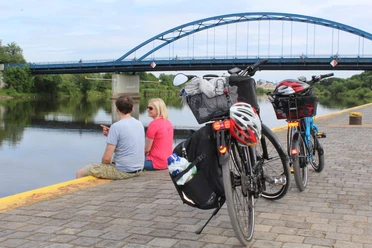 Zwei Fahrräder stehen am Ufer, während ein Paar den Ausblick auf eine Brücke über einem Fluss genießt.