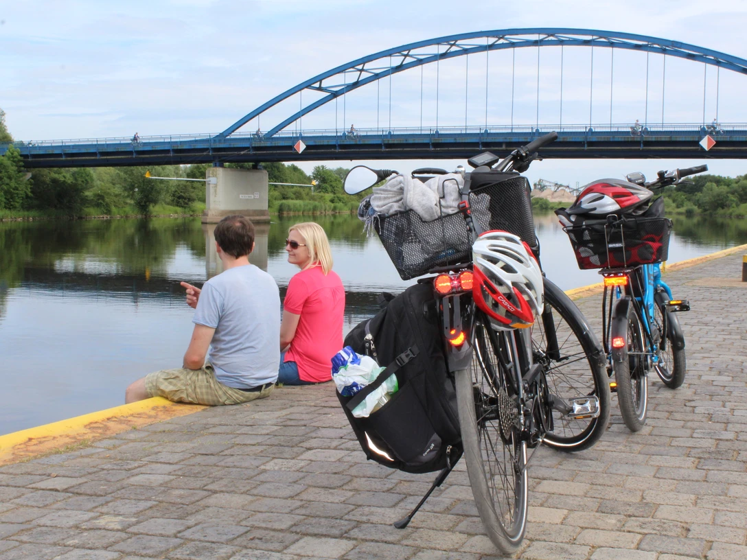 Radler Zwei Fahrräder stehen am Ufer, während ein Paar den Ausblick auf eine Brücke über einem Fluss genießt.