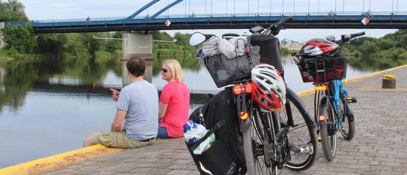 Radler Zwei Fahrräder stehen am Ufer, während ein Paar den Ausblick auf eine Brücke über einem Fluss genießt.