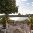 km 689 Cologne - Beach Club Sandstrand am Rheinufer mit Palmen und Sitzgelegenheiten, dominiert von einem beeindruckenden Flussblick auf den Kölner Dom und die Stadtlandschaft im Hintergrund, bei sonnigem Wetter.