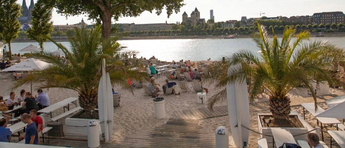 km 689 Cologne - Beach Club Sandstrand am Rheinufer mit Palmen und Sitzgelegenheiten, dominiert von einem beeindruckenden Flussblick auf den Kölner Dom und die Stadtlandschaft im Hintergrund, bei sonnigem Wetter.