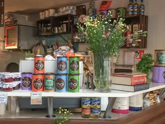 Leuchte Kaffeebar Cafétheke bestückt mit Marmeladengläsern, Blumen in einer Kristallvase, Chai-Tee Dosen und Büchern. Im Hintergrund hängen Weinkästen gefüllt mit Tassen und Tellern.Coffee bar stocked with jam jars, flowers in a crystal vase, chai tea tins, and books. Wine racks filled with cups and plates hang in the background.