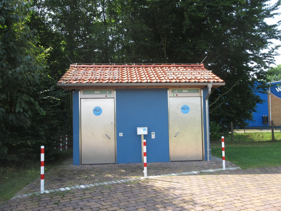 Zwei moderne, barrierefreie öffentliche Toiletten mit blauen Türen in einem parkähnlichen Umfeld.