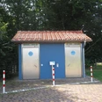 Zwei moderne, barrierefreie öffentliche Toiletten mit blauen Türen in einem parkähnlichen Umfeld.