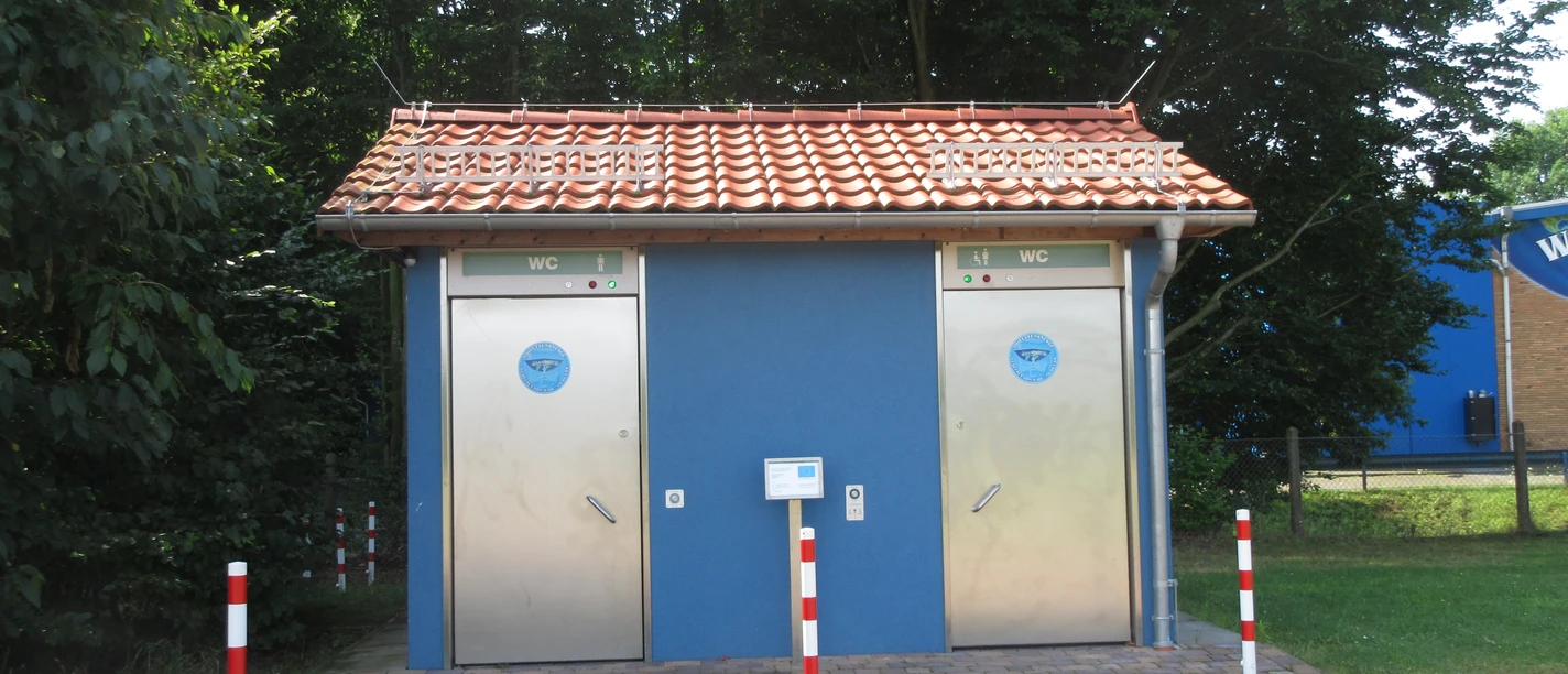 Zwei moderne, barrierefreie öffentliche Toiletten mit blauen Türen in einem parkähnlichen Umfeld.