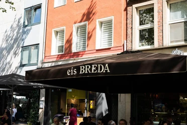 Eiscafé Breda Unter einem großen Sonnenschirm mit der Aufschrift eis Breda sitzen Menschen und essen Eis.