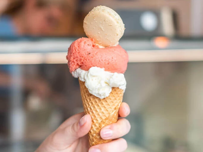 Schmelzpunkt Eine Hand hält eine Eiswaffel mit zwei Kugeln Eis, dekoriert mit einem Macaron.A hand holding an ice cream cone with two scoops of ice cream, decorated with a macaron.