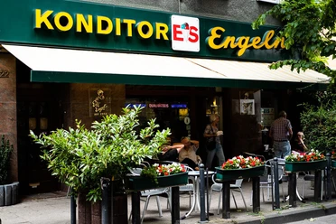 Konditorei Eis Engel Fassade mit Markise und in großen Buchstaben platziertes Schild über der Markise. Mensch sitzen vor der Konditorei.