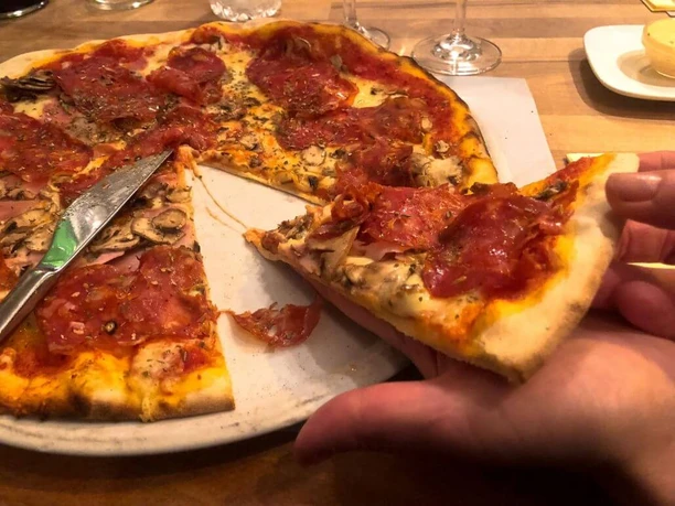 Toscanini Eine Hand hält ein Stück Pizza mit Salami und Schinken von einer angerichteten, großen Pizza mit knusprigem Rand ab.