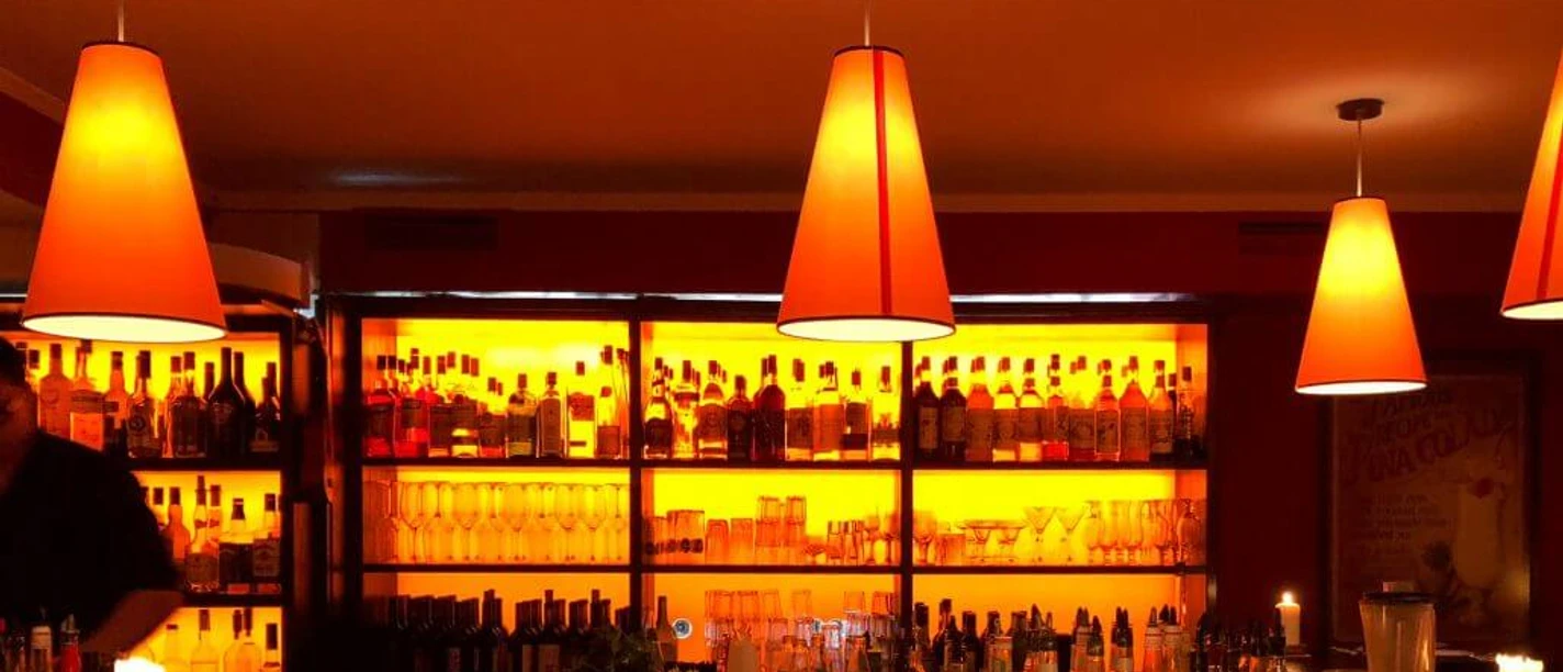 Tapeo and co The image depicts a cozy bar atmosphere illuminated by warm, orange-hued lighting emitted from stylish pendant lamps, softly highlighting the back wall filled with bottles of various beverages. Calmly flickering candles adorn the tables.
