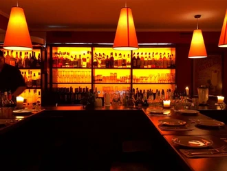 Tapeo und co Das Bild zeigt eine gemütliche Bar-Atmosphäre mit warmem orangefarbenem Licht, das von stilvollen Pendelleuchten ausgeht und die Rückwand, gefüllt mit Flaschen verschiedener Getränke, sanft erhellt. Auf den Tischen brennen ruhige Kerzen.