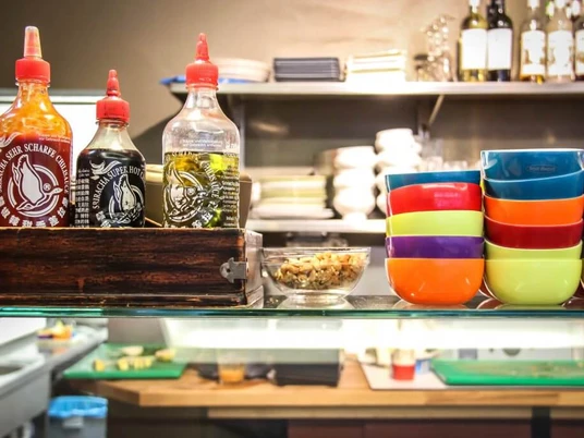 Veedel Vital Bunte Schalen stapeln sich in einem Regal einer Küche, flankiert von Flaschen mit Soßen.Colorful bowls stacked on a kitchen shelf, flanked by bottles of sauces.