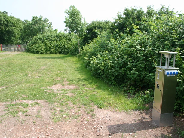 A RV parking spot in front of a green hedge and trees, with a power pole in the foreground on a gravel surface.