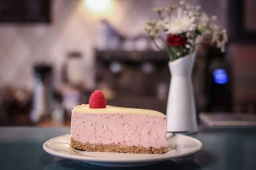 Wo ist Tom? Ein Stück Himbeer-Käsekuchen mit einer frischen Himbeere als Dekoration auf einem Teller. Im Hintergrund sind verschwommen eine Blumenvase und eine Kaffeemaschine zu erkennen.