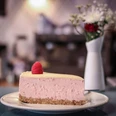 Wo ist Tom? Ein Stück Himbeer-Käsekuchen mit einer frischen Himbeere als Dekoration auf einem Teller. Im Hintergrund sind verschwommen eine Blumenvase und eine Kaffeemaschine zu erkennen.A slice of raspberry cheesecake decorated with a fresh raspberry, served on a plate. A blurred flower vase and coffee machine can be seen in the background.