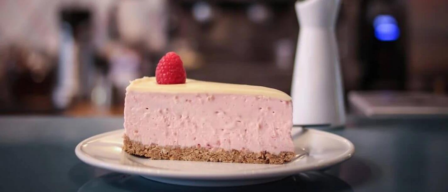 Wo ist Tom? Ein Stück Himbeer-Käsekuchen mit einer frischen Himbeere als Dekoration auf einem Teller. Im Hintergrund sind verschwommen eine Blumenvase und eine Kaffeemaschine zu erkennen.