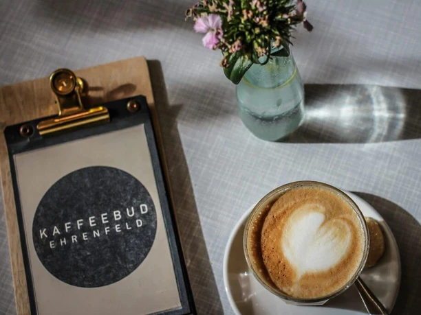 KAFFEEBUD Nahaufnahme, ein Cappuccino, daneben die Menükarte vom Café und in der Mitte eine Vase mit Blumen.