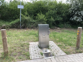 Öffentlicher Wohnmobil-Stellplatz mit Edelstahl-Säule zur Ver- und Entsorgung nahe einer grünen Hecke.