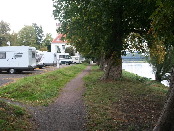 Wohnmobilstellplatz an einem Flussufer mit Bäumen und geparkten Wohnmobilen entlang eines Schotterwegs.
