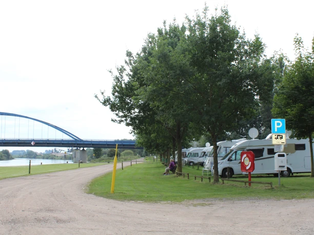 Wohnmobilstellplatz am Weserradweg mit Blick auf eine Brücke; Bäume bieten Schatten und Erholung.