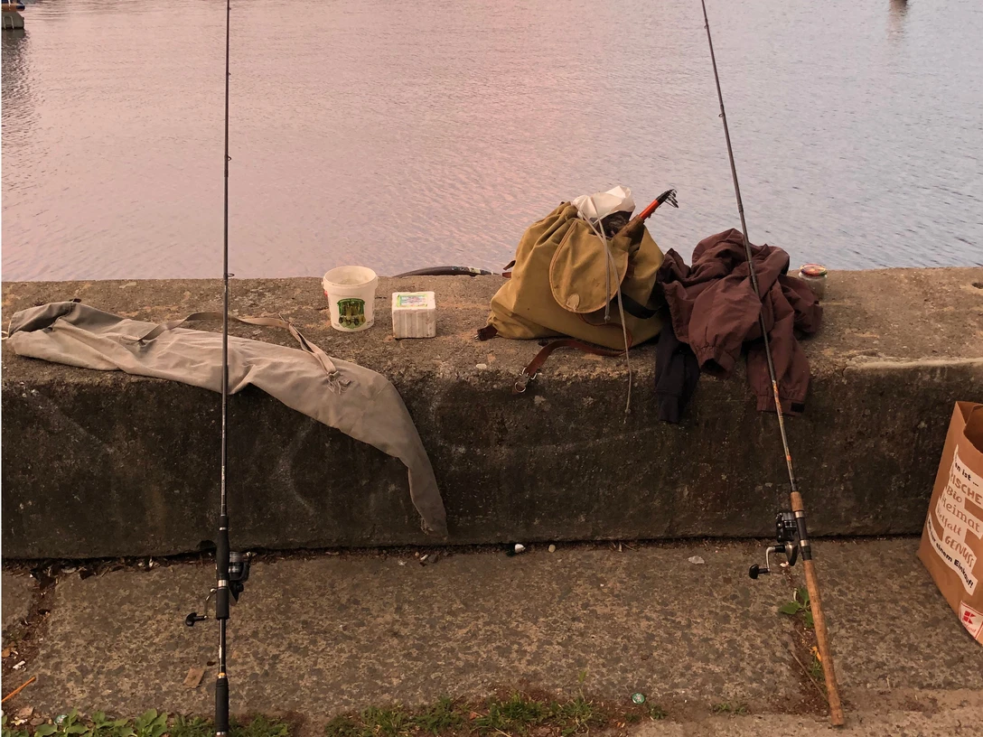 Zwei Angelruten ruhen auf einer Betonmauer am Ufer eines Sees, umgeben von Angelausrüstung.