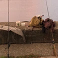 Zwei Angelruten ruhen auf einer Betonmauer am Ufer eines Sees, umgeben von Angelausrüstung.