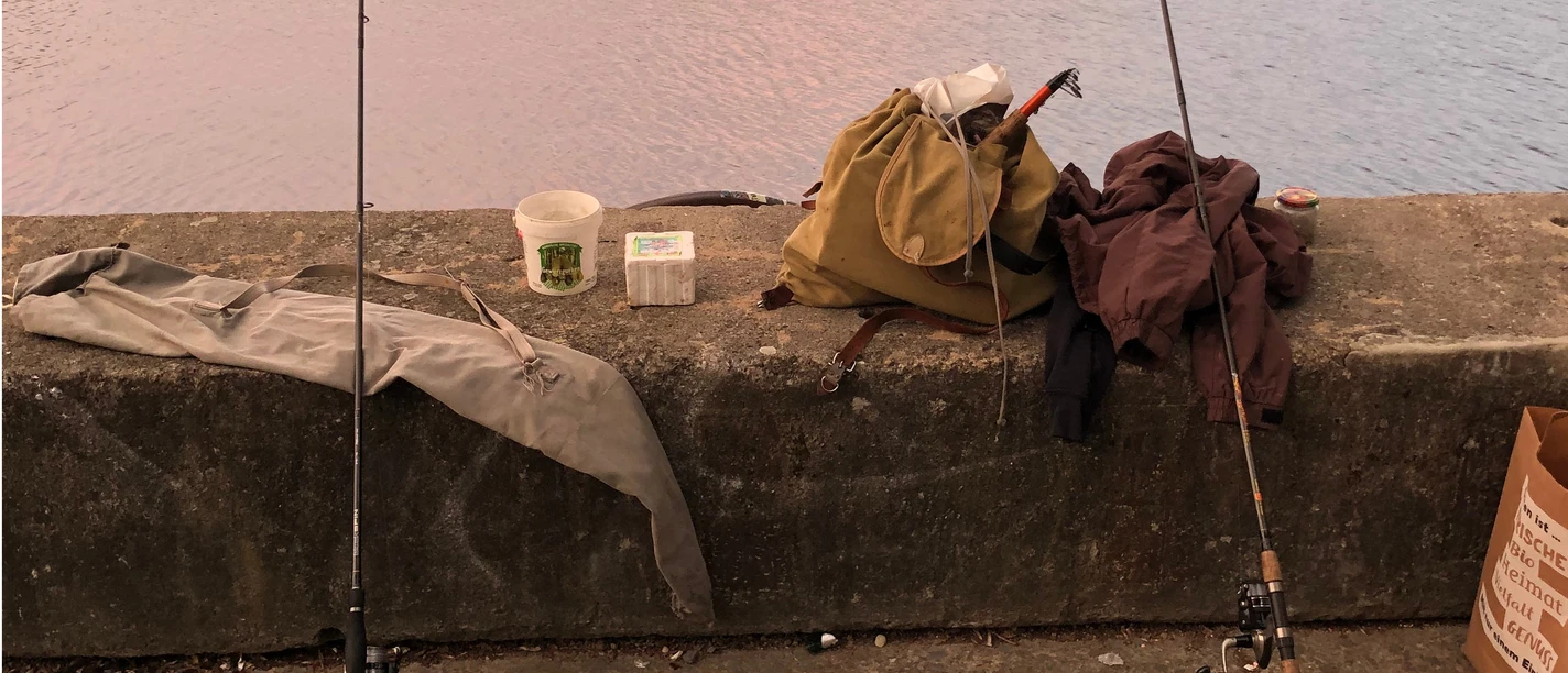 Angeln am Estorfer See Zwei Angelruten ruhen auf einer Betonmauer am Ufer eines Sees, umgeben von Angelausrüstung.