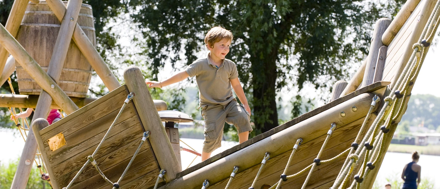 Freizeitanlage - Schiff Kind spielt auf hölzernem Kletterschiff am See, umgeben von Bäumen und sommerlicher Atmosphäre.