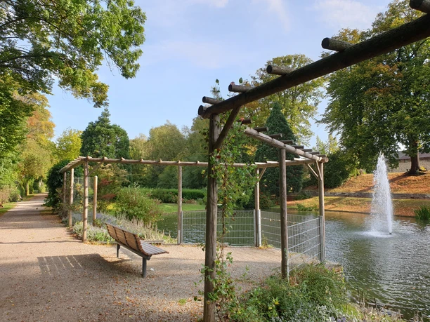 Bankje onder een houten pergola bij de vijver met fontein in het groene Bäckerwall park in Einbeck.