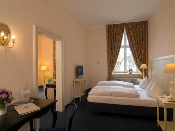 Helle Juniorsuite im ersten Stock mit Doppelbett, Sessel, Schreibtisch, TV und großen Fenstern.